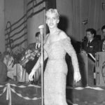 A young blonde woman in a tight dress sings in front of a bandstand