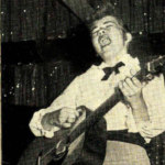 A white woman with bangs styled to look like Elvis's bouffant hairdo plays guitar. Her eyes are closed.