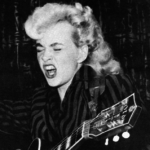 A young blonde woman with a guitar. Here eyes are closed and she appears to be singing loudly,