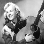 A young white woman with light curly hair posed with an acoustic guitar. She is wearing a western-style fringed shirt.