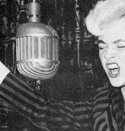 A young woman with blond hair sings into an old fashioned microphone with her eyes shut, as if she is singing loudly
