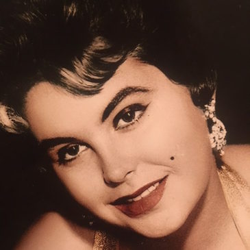 A cropped headshot of a white woman with black hair and heavy makeup, wearing dangly earrings